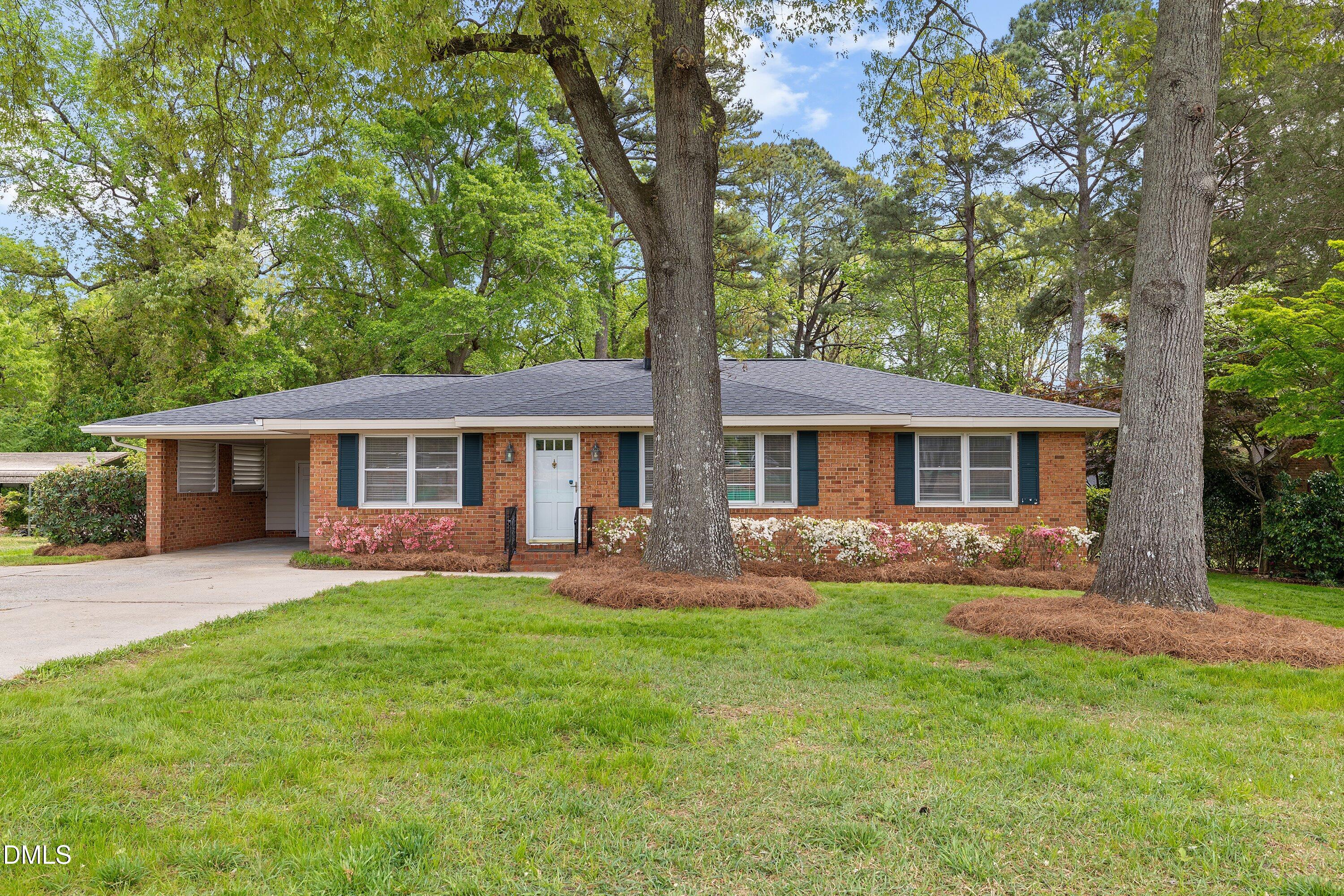 Red brick ranch home with a covered carport, flowering shrubs, and mature trees across the lawn.