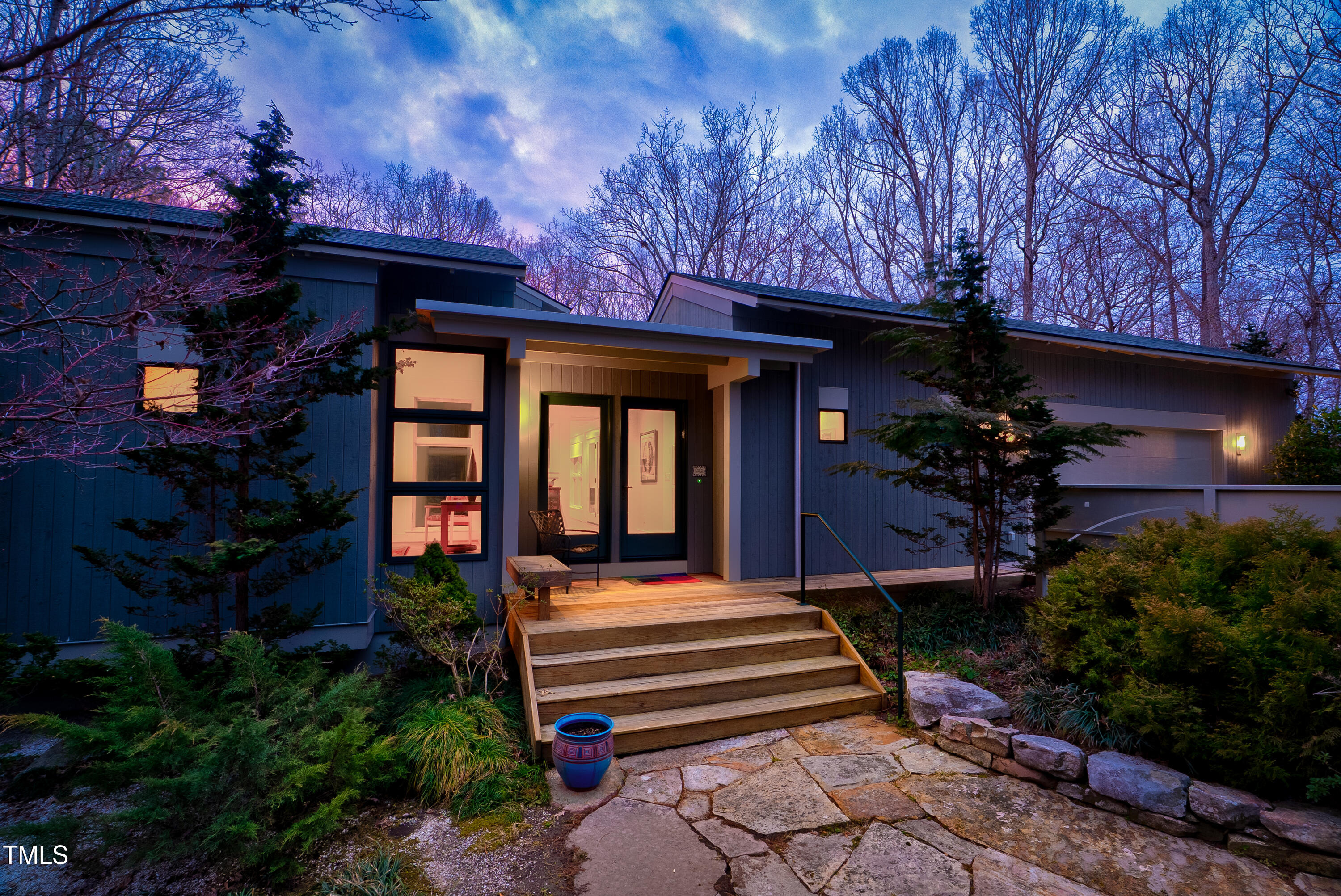 Modern blue-sided house with a warmly lit entry, broad wooden steps, and a stone path at dusk.