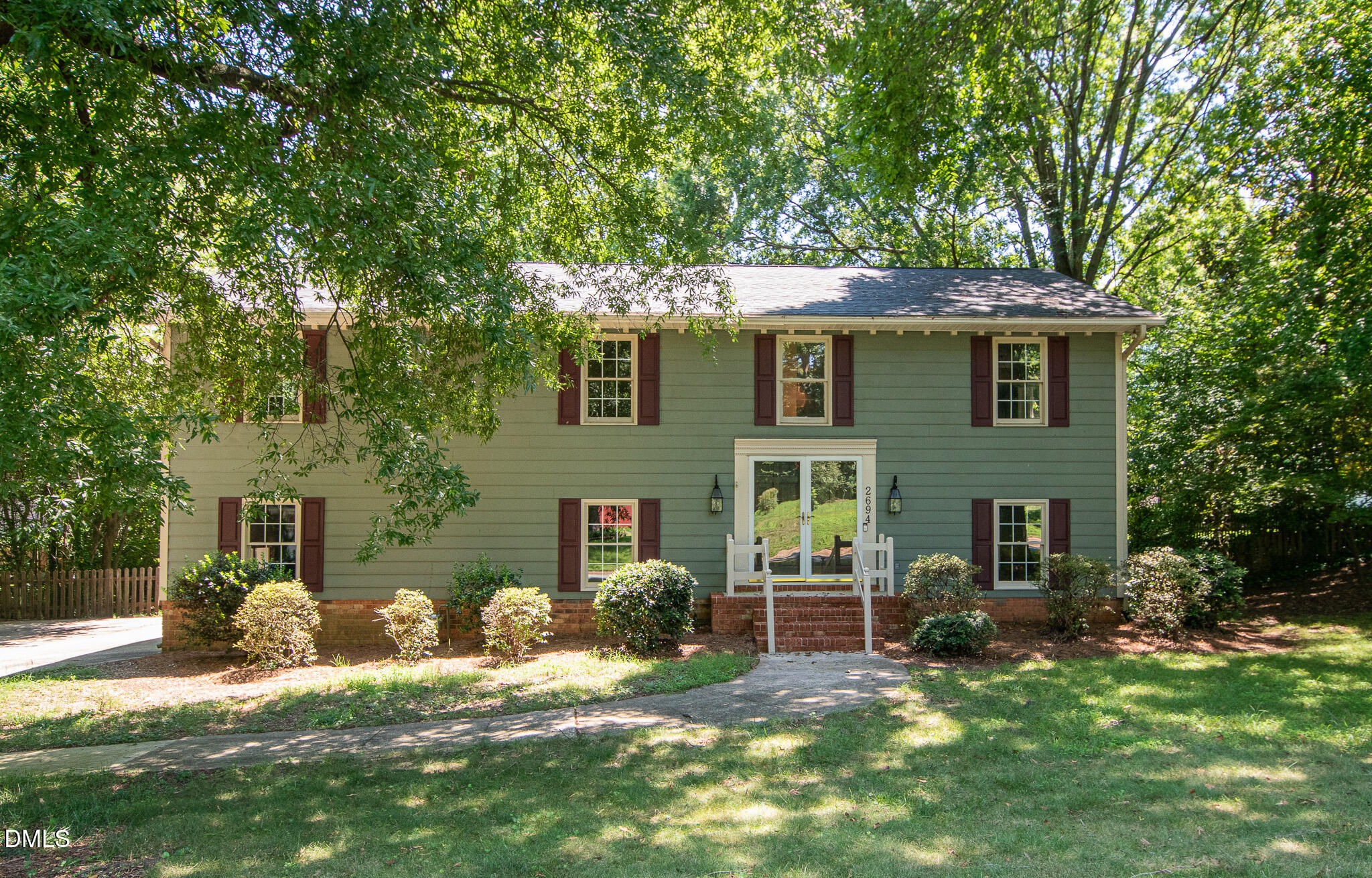 Green two-story house with red shutters, brick foundation, and mature trees shading the front lawn.