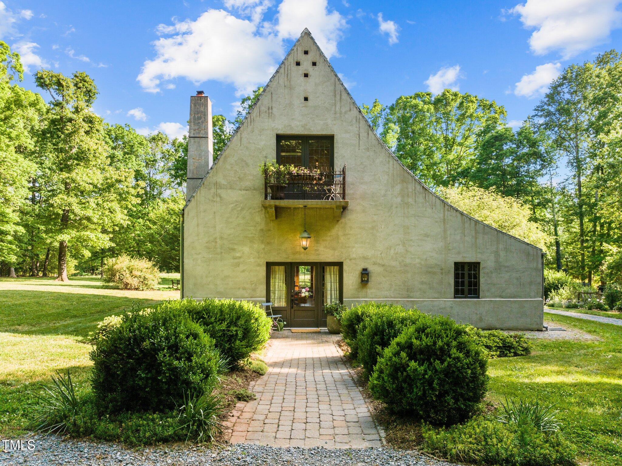 Stucco house with a steep triangular facade and balcony, approached by a brick walkway through landscaped grounds.