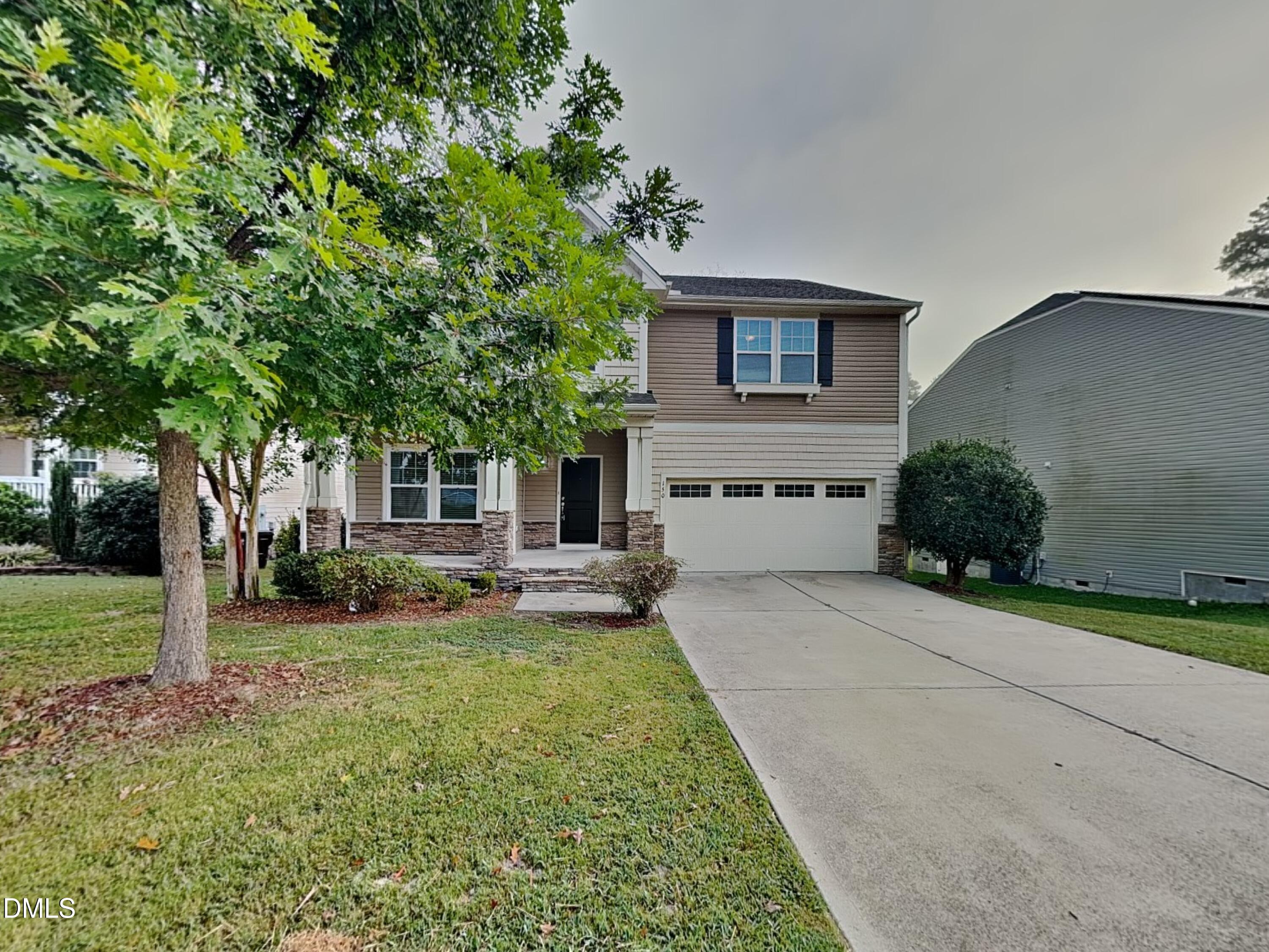 Two-story beige house with stone accents, a two-car garage, and a tree-shaded front lawn.