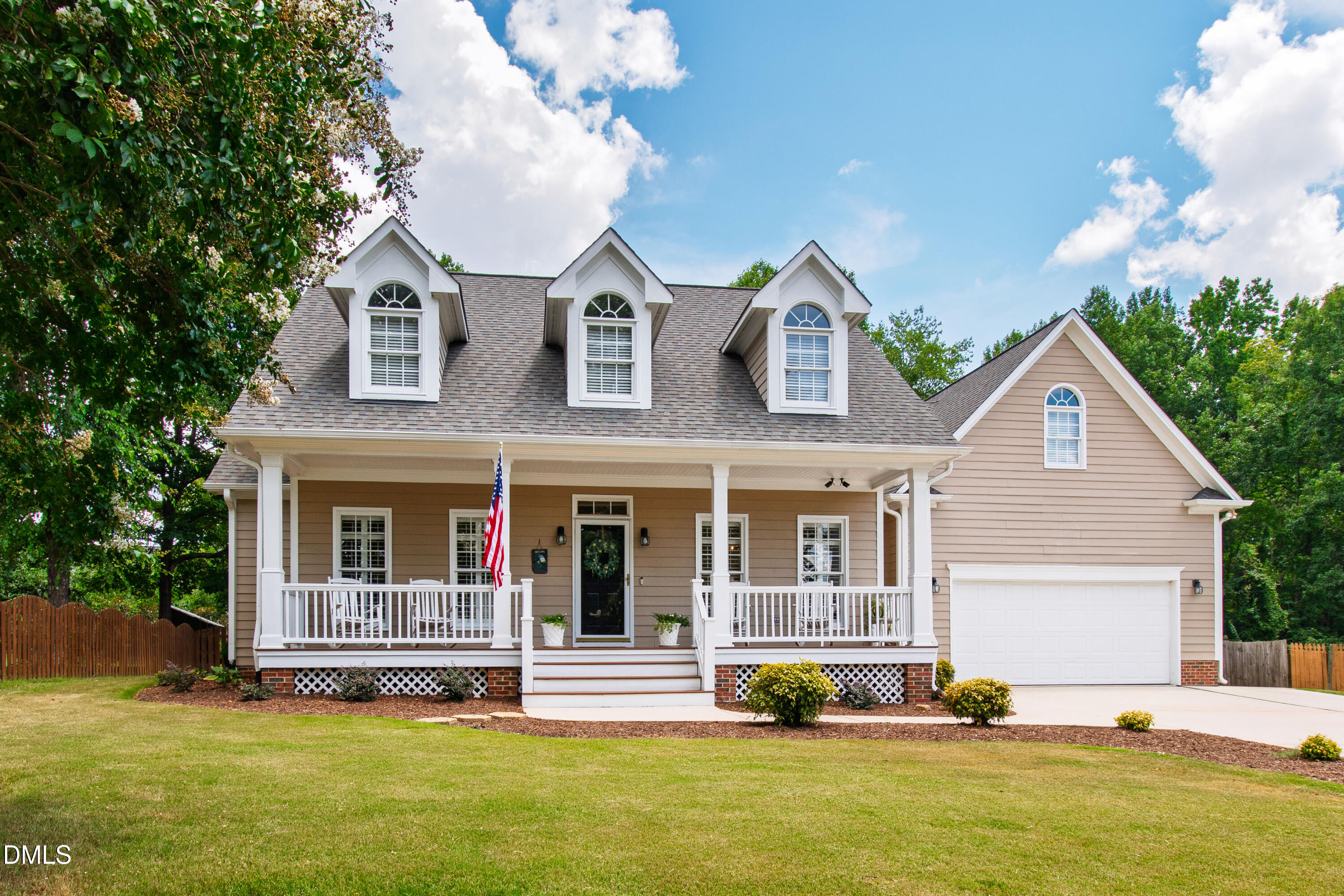 Two-story beige house with a white wraparound porch, three dormers, and an attached garage.