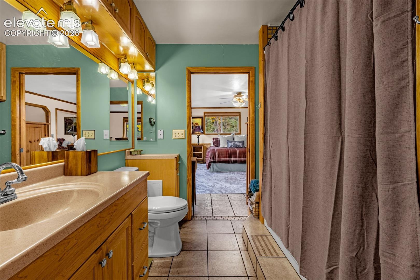 Bathroom featuring a wood vanity with a solid-surface integrated sink, a full-width mirror with integrated lighting, and ceramic tile flooring