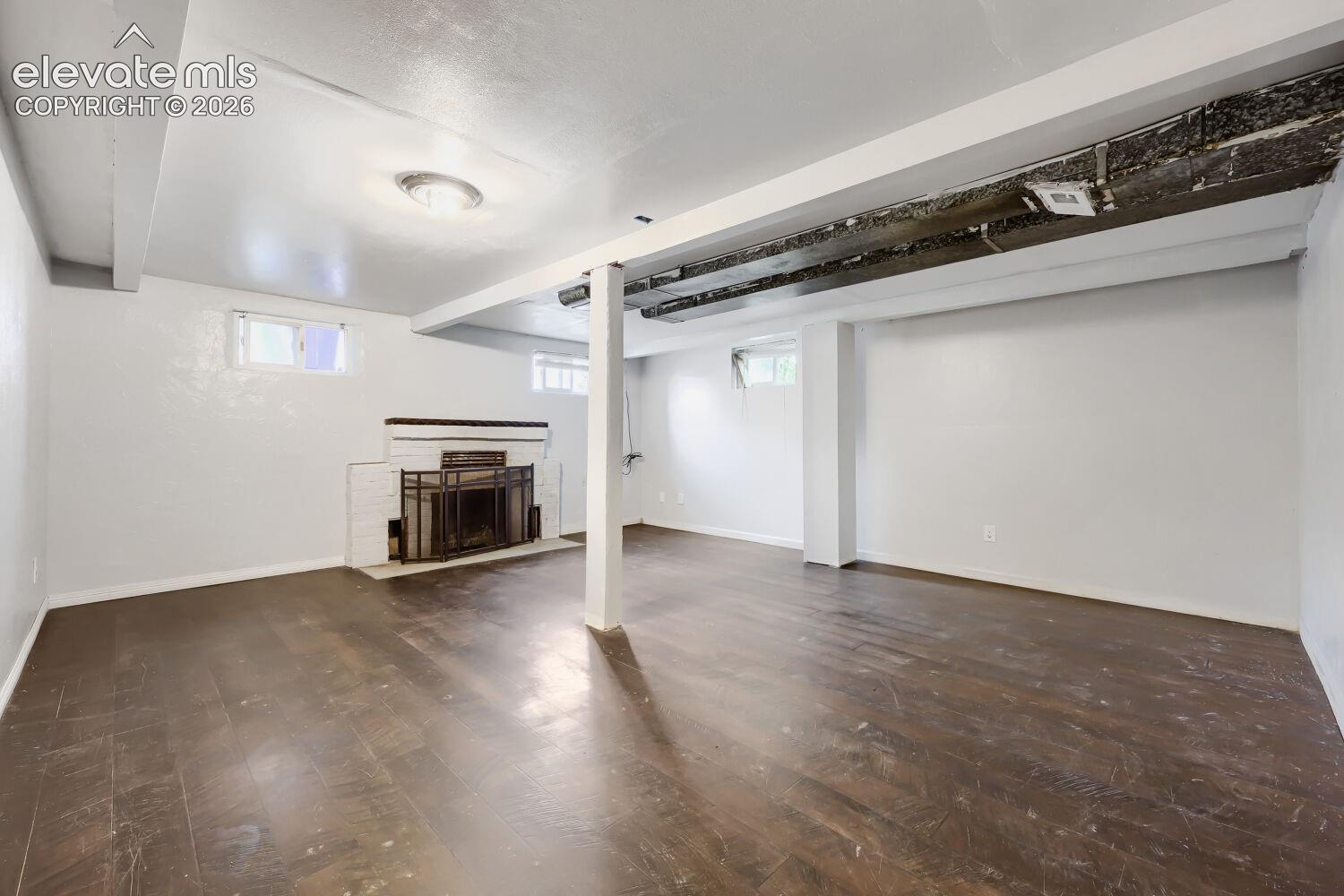 Expansive room featuring wood-finish flooring, white painted walls, exposed ceiling beams, and a fireplace with a black metal screen