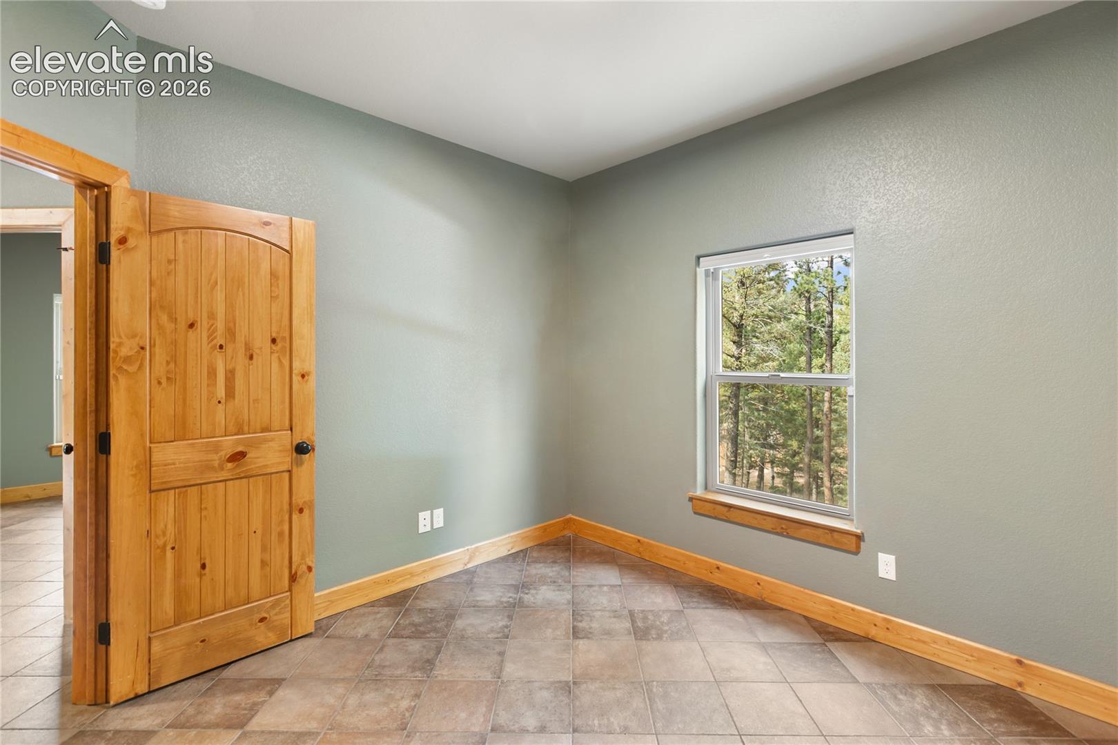 Room interior featuring tile flooring, natural wood door and trim, and single-hung window with exterior forest backdrop