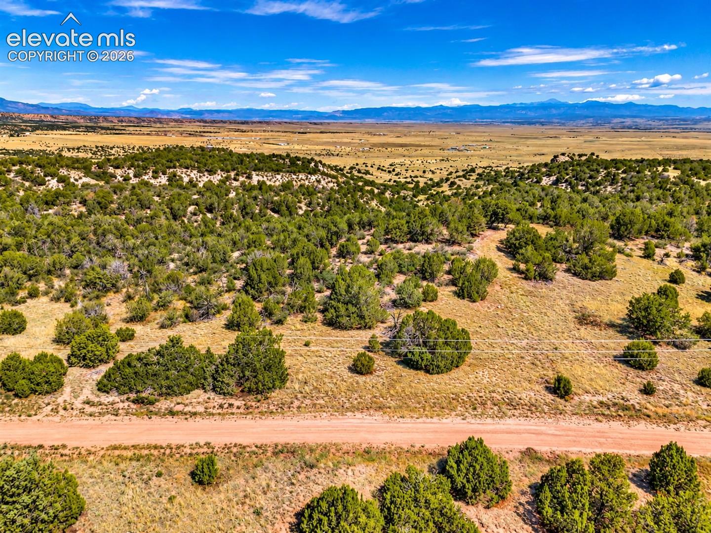Aerial perspective showcasing the rural roadway frontage, rolling juniper-covered hills, and sweeping views across the open valley toward distant mountain peaks.

