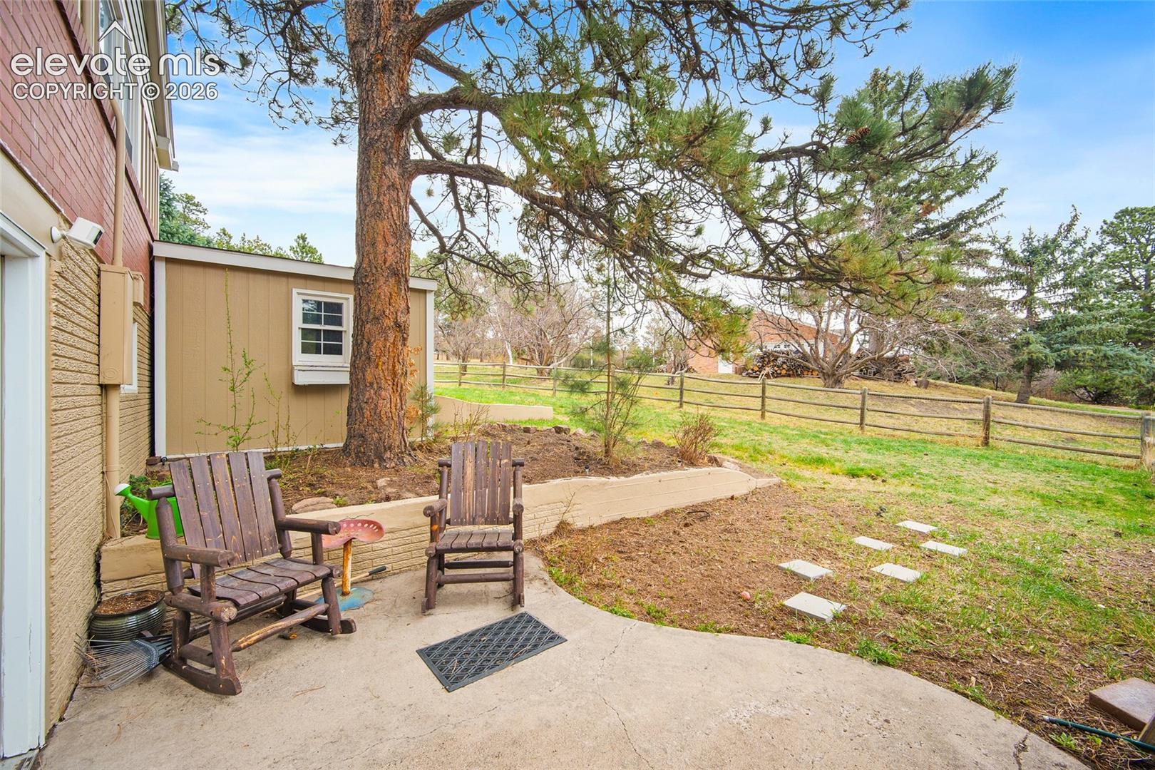 Concrete patio featuring a low-profile stone retaining wall, mature trees, and a post-and-rail perimeter fence