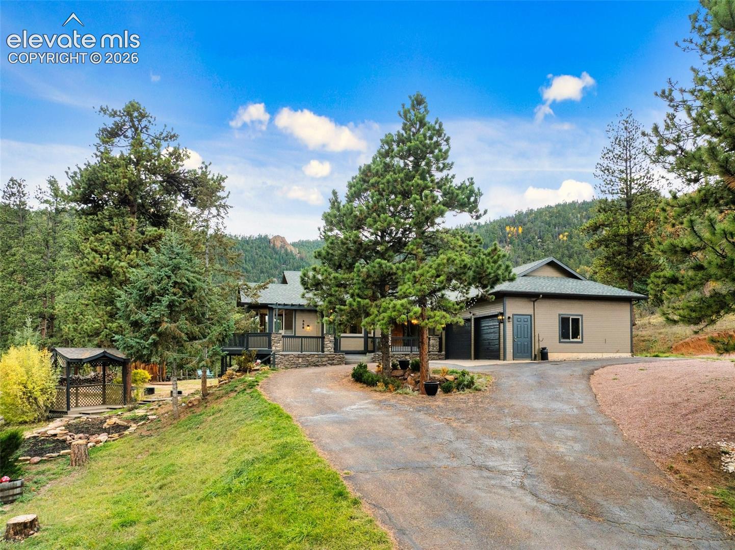 Exterior featuring multi-gabled rooflines, stone foundation detailing, asphalt driveway, attached garage, and surrounding mature conifer trees