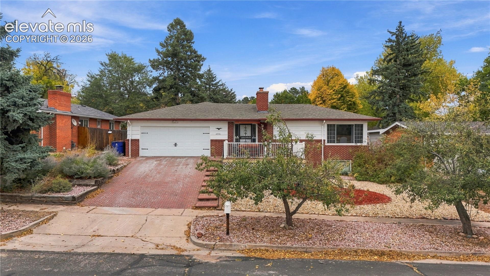 Single-story brick and siding exterior featuring a multi-car garage, front porch with metal railing, brick chimney, and mature landscaping