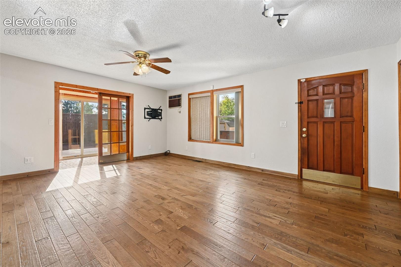 Living area featuring wood-finish flooring, a wood-frame glass sliding door, a solid wood entry door with glass paneling, a multi-blade ceiling fan, and multi-pane windows