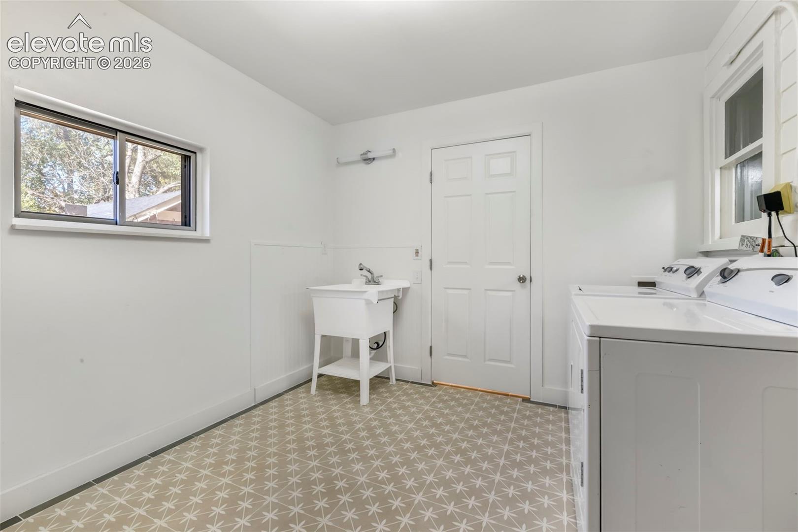 Laundry area featuring geometric patterned tile flooring, a utility sink with a chrome faucet, dual windows, and a six-panel door