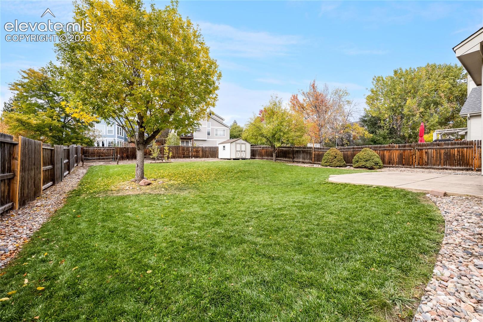Fenced backyard featuring a concrete patio, detached storage shed, mature trees, and rock landscape borders