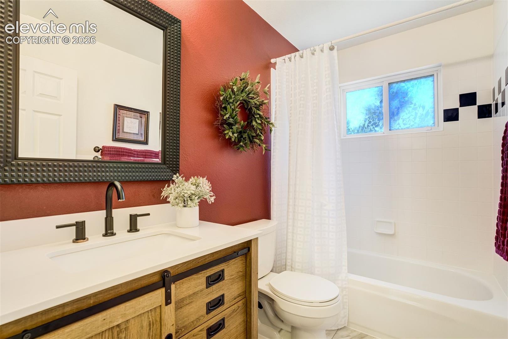 Full Bathroom main levelfeaturing a wood-finish vanity with a sliding barn-door-style cabinet, a white countertop with an integrated basin, a matte black widespread faucet, a textured framed wall mirror, and a tiled tub-shower combination