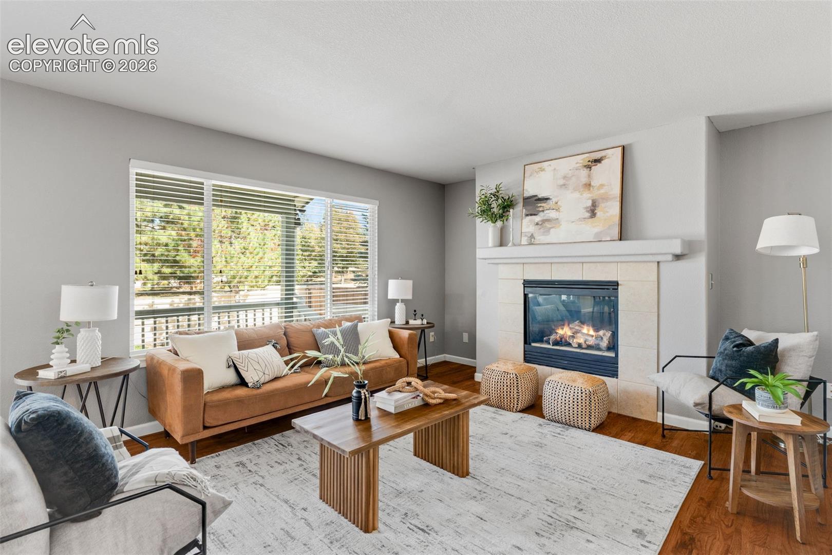 Living area featuring large windows with horizontal blinds, wood-finish flooring, and a fireplace with a light-toned tile surround and mantle