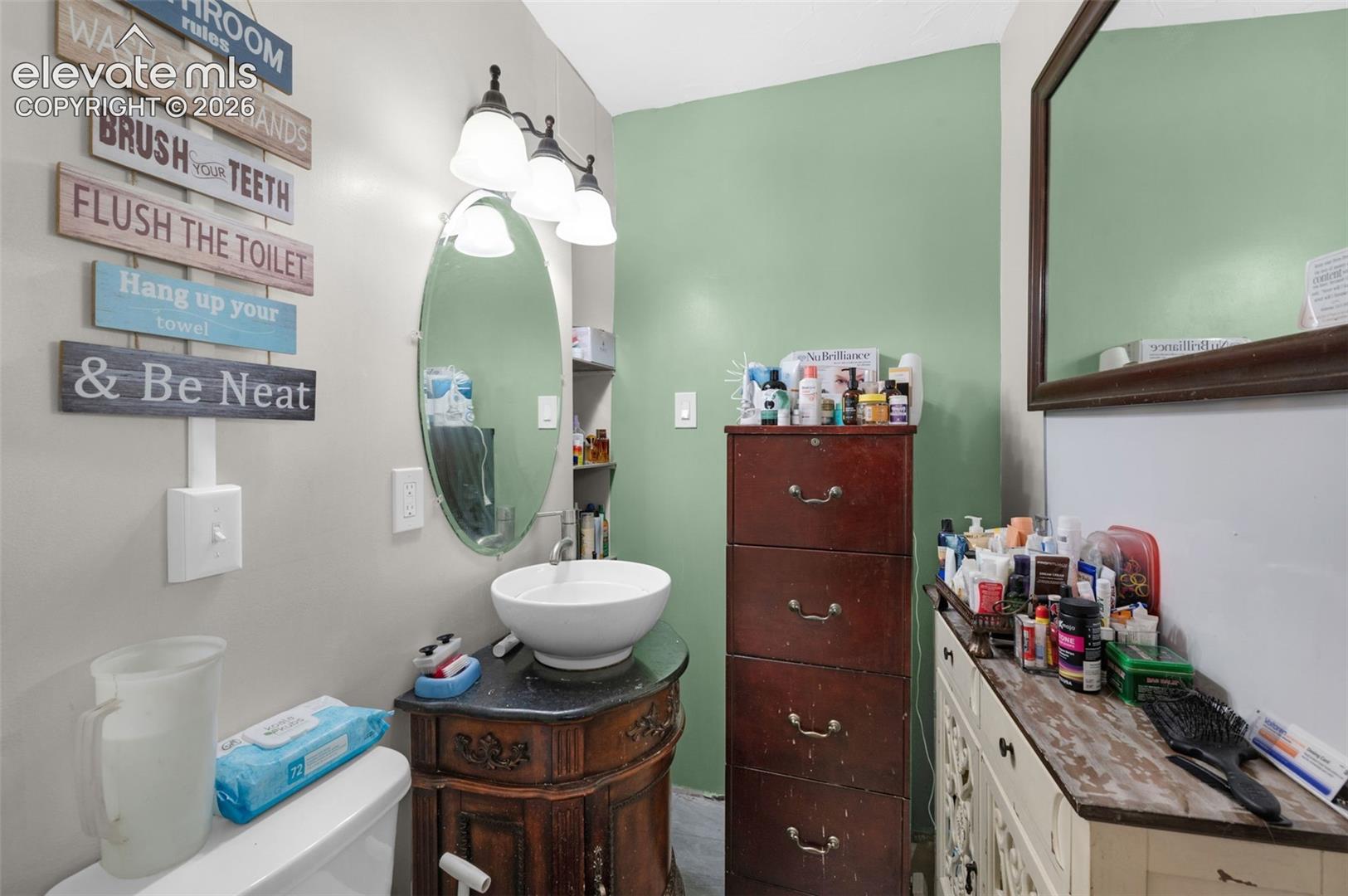Bathroom featuring a vessel sink with a dark stone-finish countertop, a wooden vanity base with decorative molding, and an oval wall-mounted mirror with an integrated three-light fixture