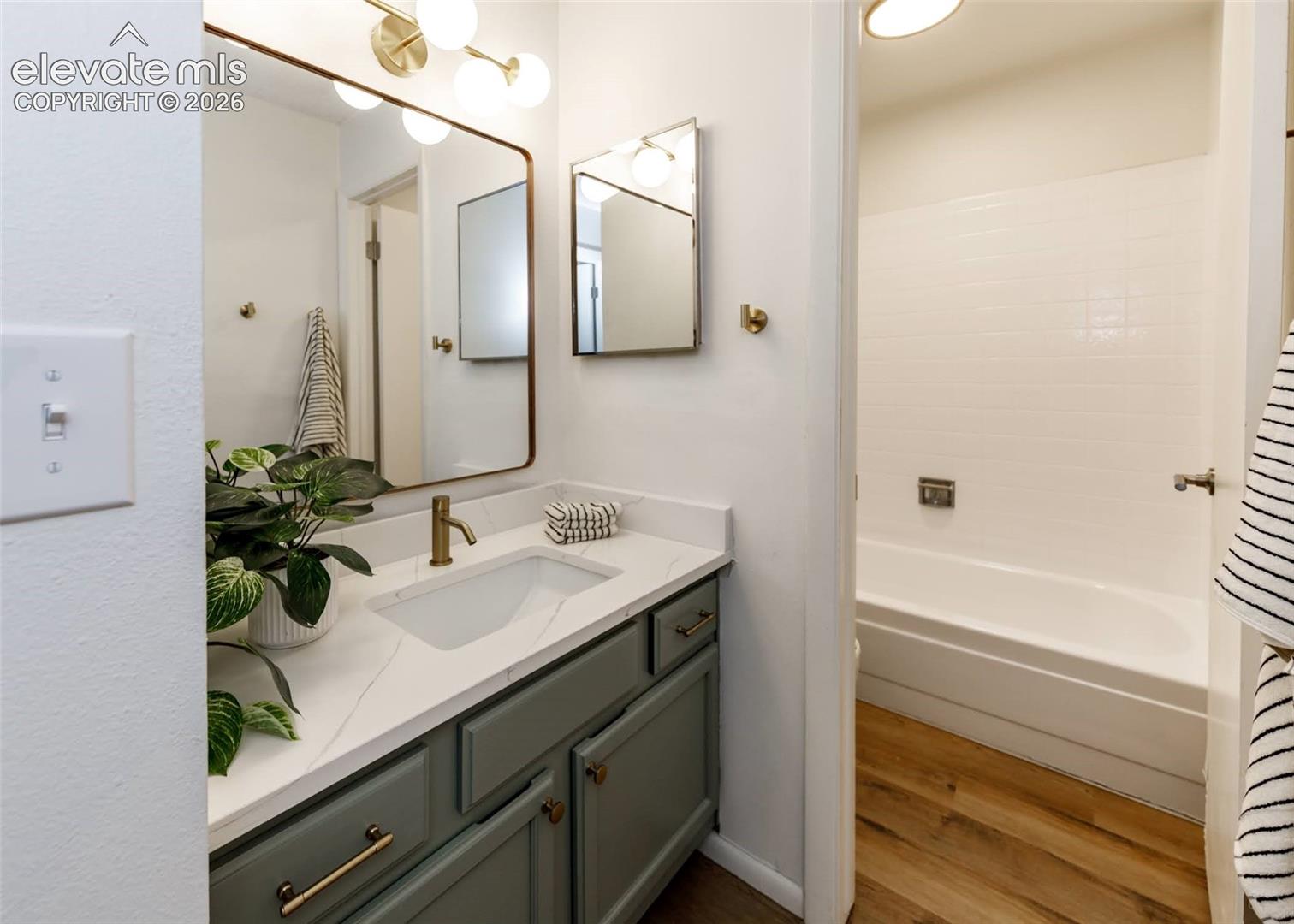 Bathroom featuring a custom-colored vanity, quartz countertop, brass-finish faucet, rectangular drop-in sink, and wood-finish flooring