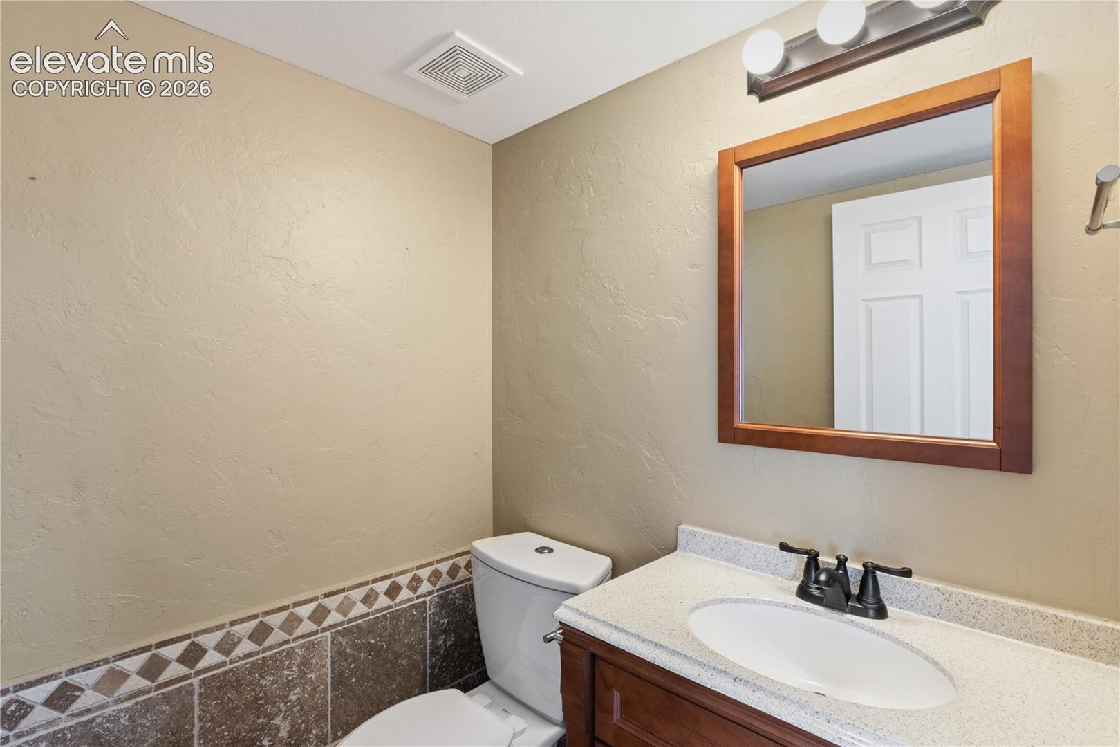 Vanity featuring a stone-composite countertop, undermount oval sink, and dual-handle matte black faucet, complemented by a framed wall mirror, three-light vanity fixture, and tiled wainscoting with a decorative diamond accent border