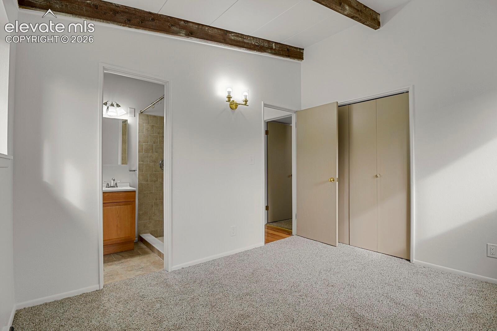 Interior room featuring vaulted ceilings with exposed wood beams, wall-to-wall carpeting, dual-door closet storage, and a wall-mounted brass light fixture looking into the attached 3/4 bath
