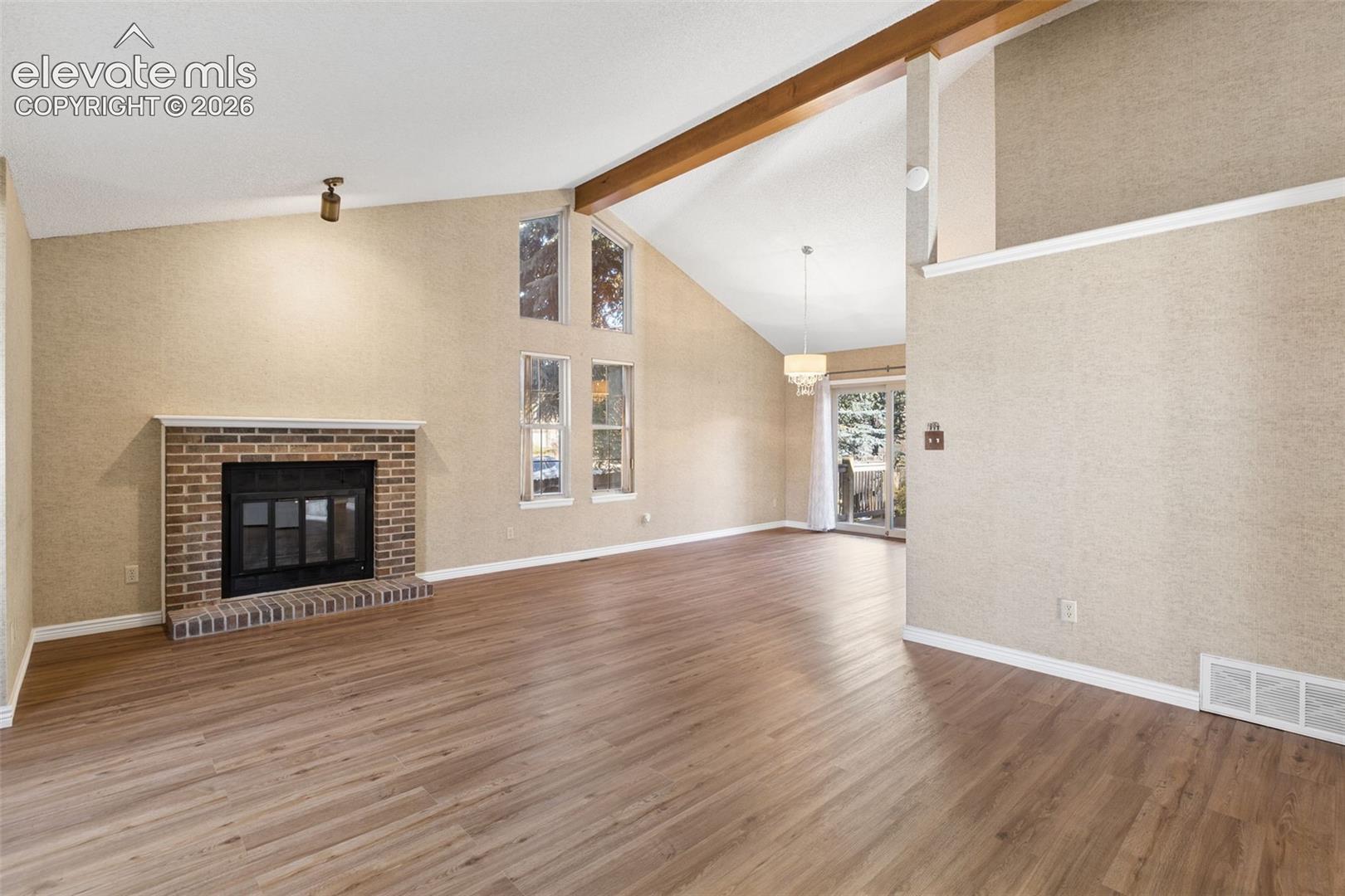 Vaulted ceilings featuring exposed wood-finish beams, brick fireplace with a wood-finish mantel, wood-finish flooring, and vertical multi-pane windows