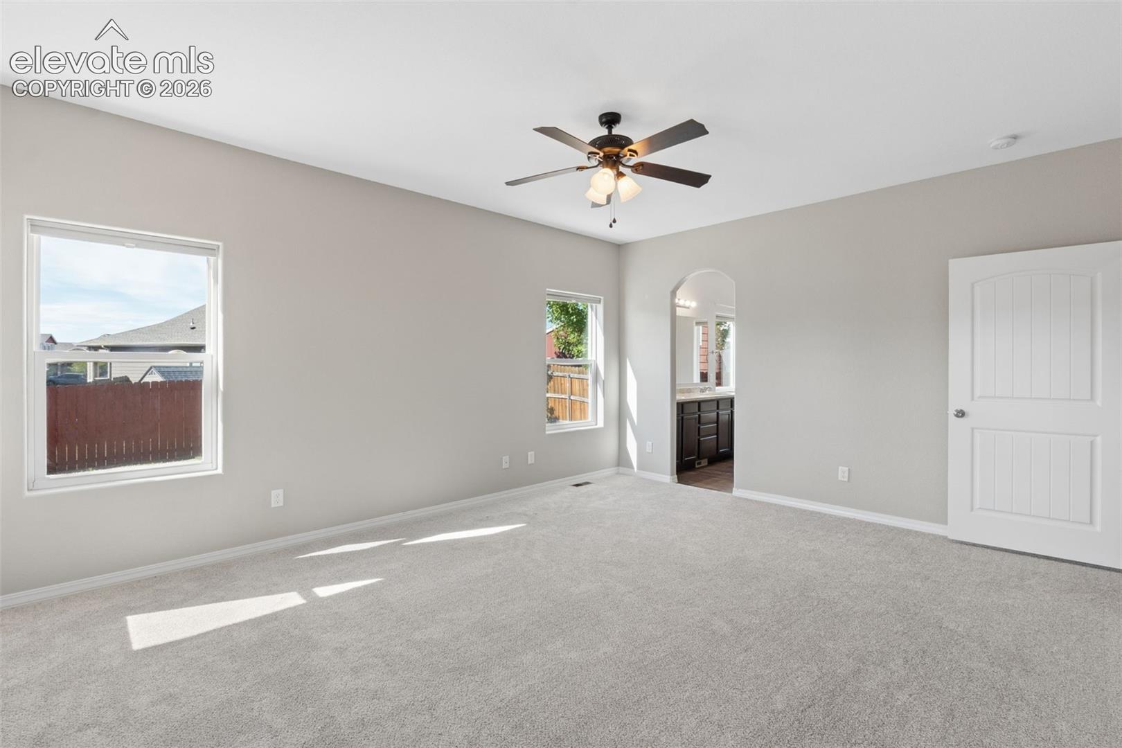 Spacious room featuring light gray walls, plush gray carpeting, two windows providing natural light, and an arched entryway leading to a separate area with dark wood cabinetry