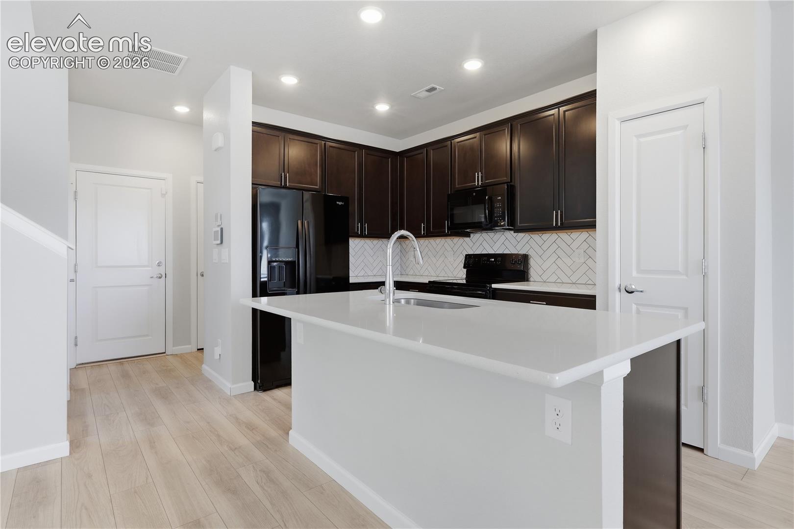 Modern kitchen featuring a light-toned island with an integrated sink, dark wood-finish cabinetry, black appliances, a white subway tile backsplash, and recessed lighting