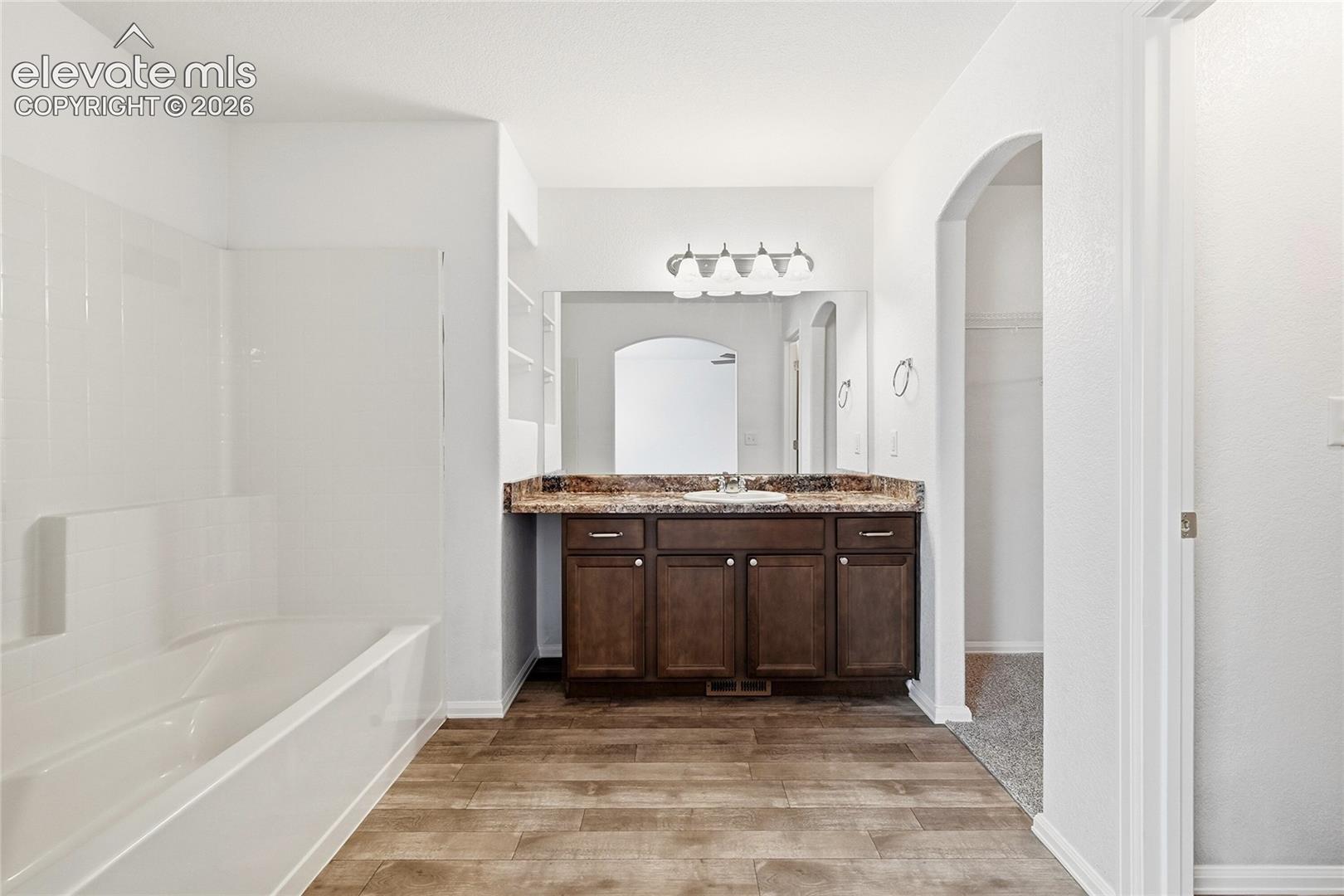 Bathroom featuring a dark wood-finish vanity, stone-patterned countertop with an integrated sink, full-width vanity mirror, four-light wall-mounted fixture, and wood-finish flooring