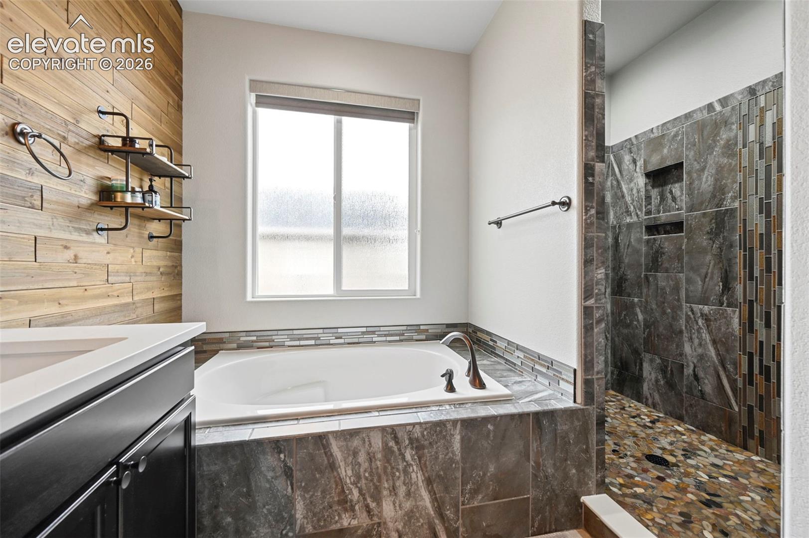 Bathroom featuring wood-plank accent wall, soaking tub with tile surround, walk-in shower with stone tile walls, pebble-style shower flooring, and built-in wall shelving