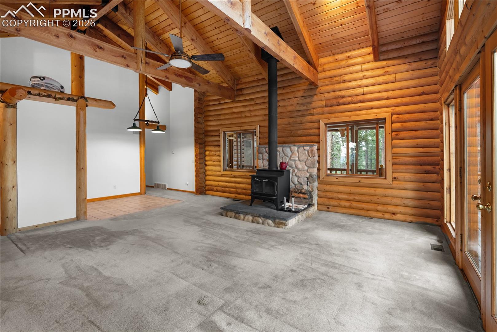 Unfurnished living room with a wood stove, a wooden ceiling with exposed beams, carpet floors, a ceiling fan, and rustic walls