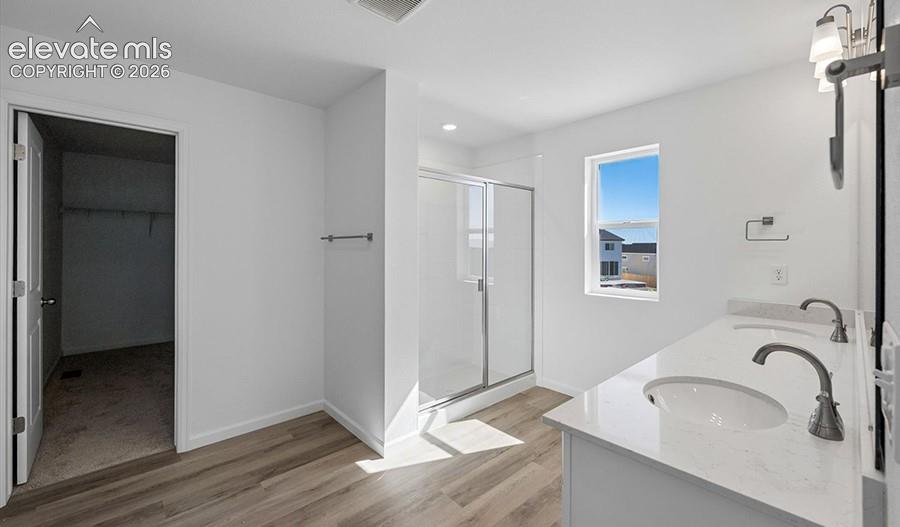 Dual-sink vanity with a solid-surface countertop and metal fixtures, wood-finish flooring, glass-enclosed shower, and built-in walk-in closet