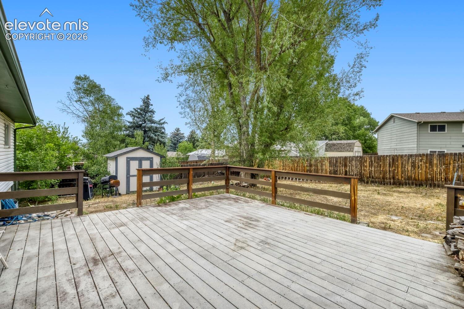 Spacious outdoor deck with wood railing, featuring a large backyard, mature trees, and a storage shed