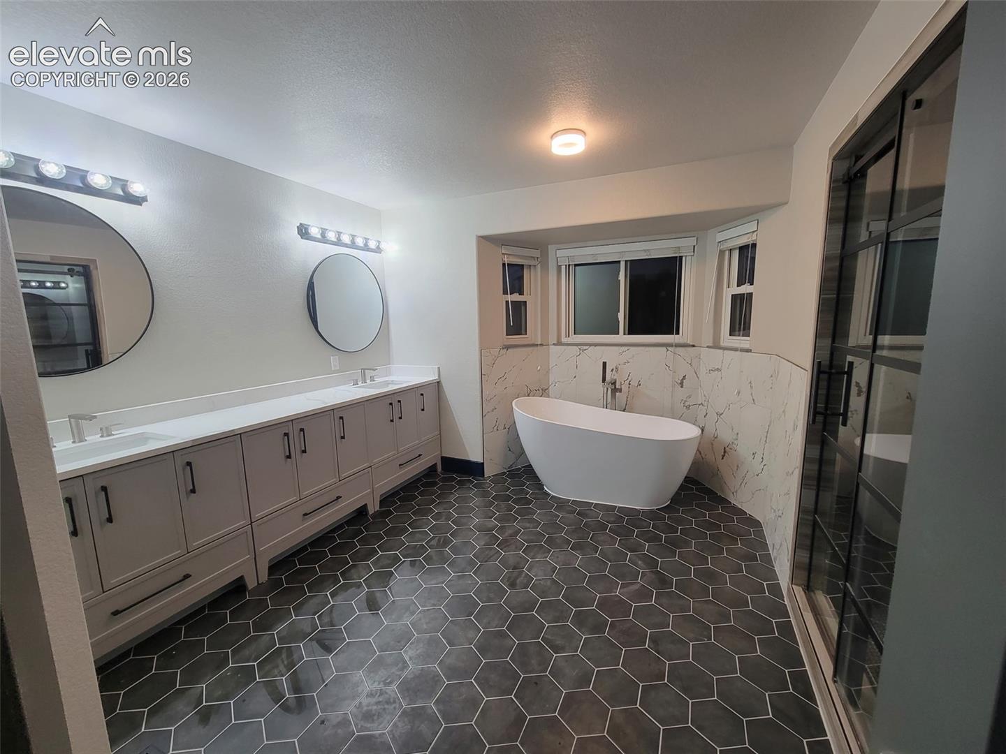 Dual vanity with undermount sinks, hexagonal floor tiling, freestanding soaking tub, and a glass-enclosed shower