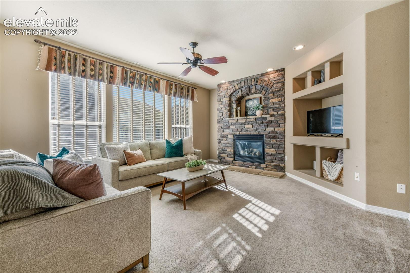 Stone-faced fireplace with hearth, built-in shelving, recessed lighting, ceiling fan, and large windows with blinds