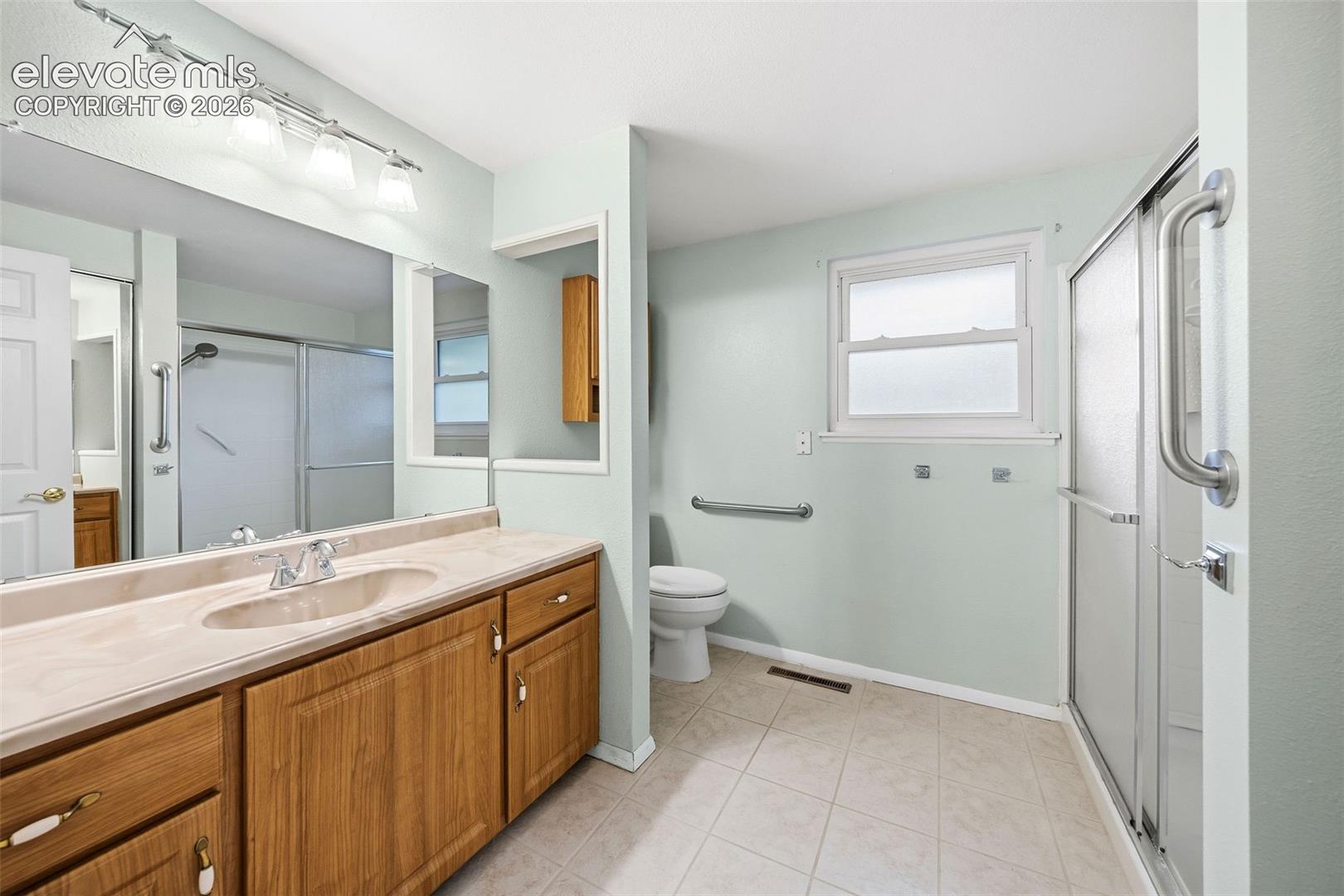 Bathroom featuring wood-finish cabinetry, a large wall-to-wall mirror, integrated vanity countertop, ceramic tile flooring, and a glass-enclosed shower