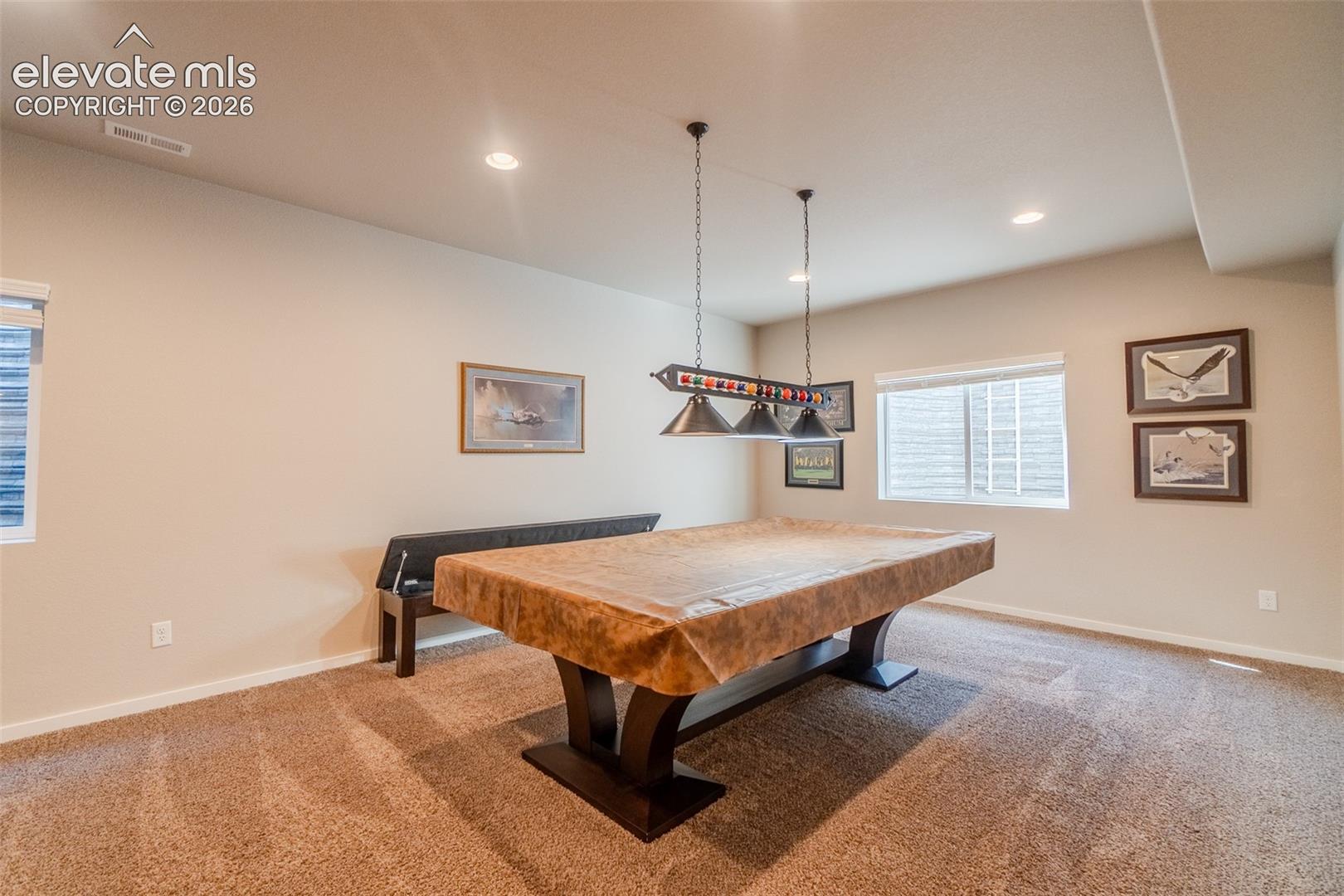 Recessed lighting illuminates the carpeted room, featuring a window with horizontal blinds and a pool table with a dedicated overhead light fixture
