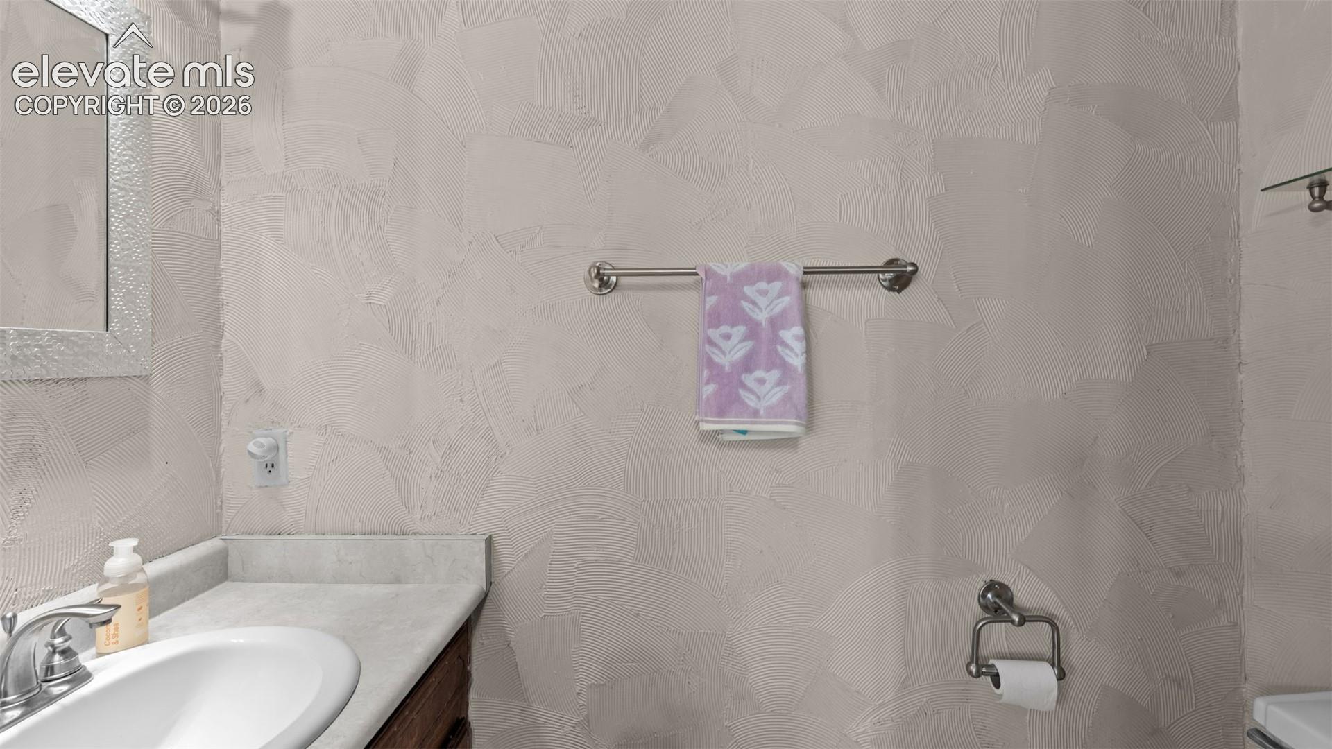 Textured wall finish featuring a vanity with a stone-surface countertop, integrated basin, brushed metal faucet, and wall-mounted metal towel bar