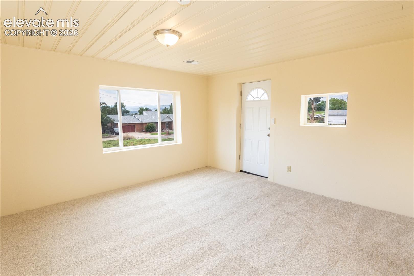 Interior space featuring neutral wall-to-wall carpeting, beadboard ceiling, flush-mount lighting, large picture window, and exterior entry door with an arched glass inset