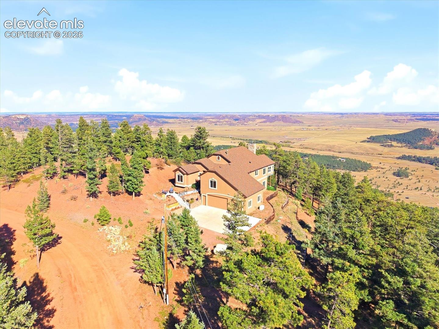 Aerial perspective of a multi-level residence featuring a multi-pitched shingle roof, attached garage, concrete driveway, and expansive hillside acreage with native evergreen trees