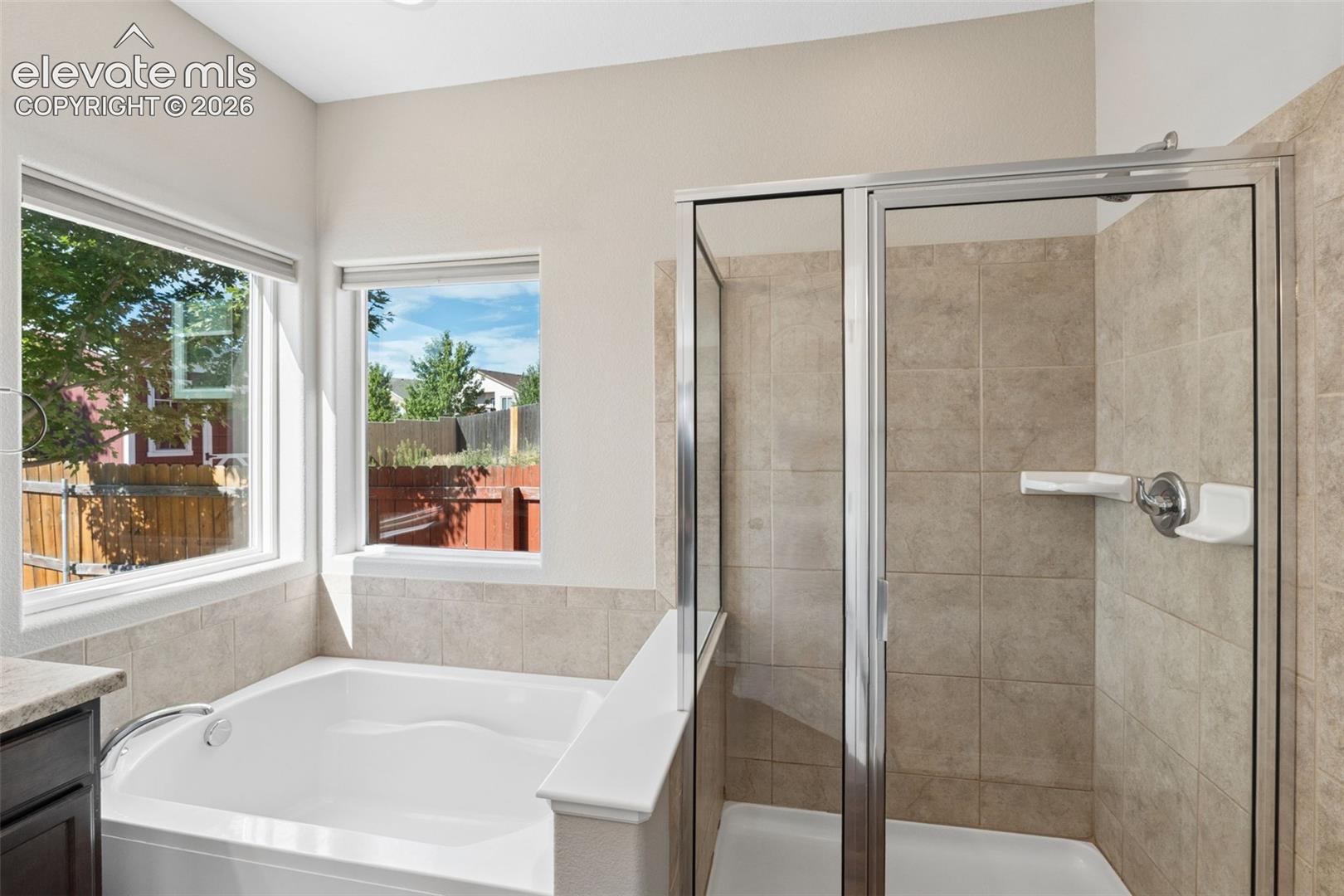 Spacious bathroom featuring a built-in soaking tub with surrounding windows, a glass-enclosed shower with tile surround, and a dark wood-finish vanity with a stone-finish countertop