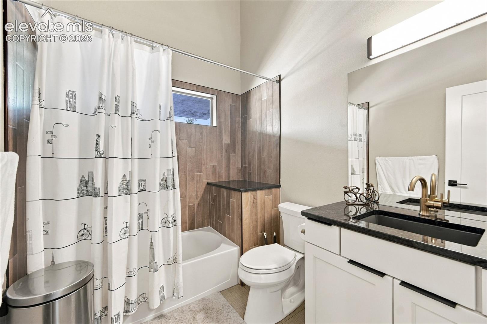 Bathroom featuring a vanity with dark stone countertop, an integrated rectangular sink, wall-to-wall mirror, and wood-finish tile surround in the tub area with vaulted ceilings