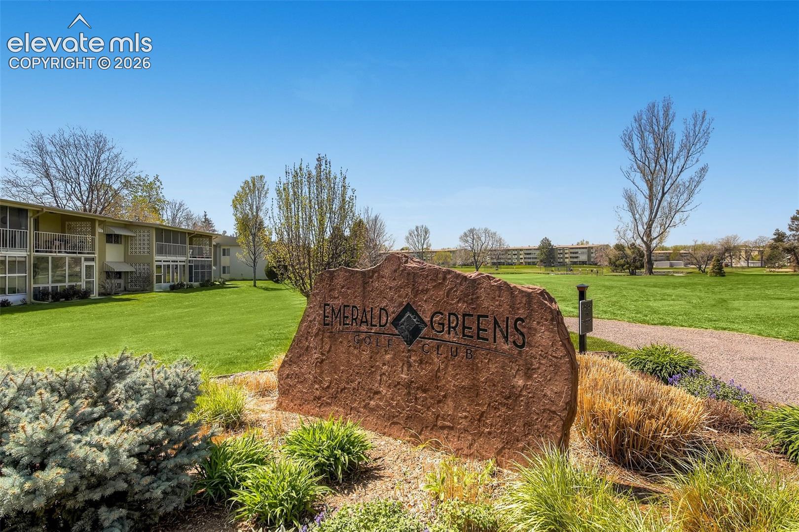 Property entrance featuring a stone monument sign, landscaped beds, a paved pathway, and a large green space