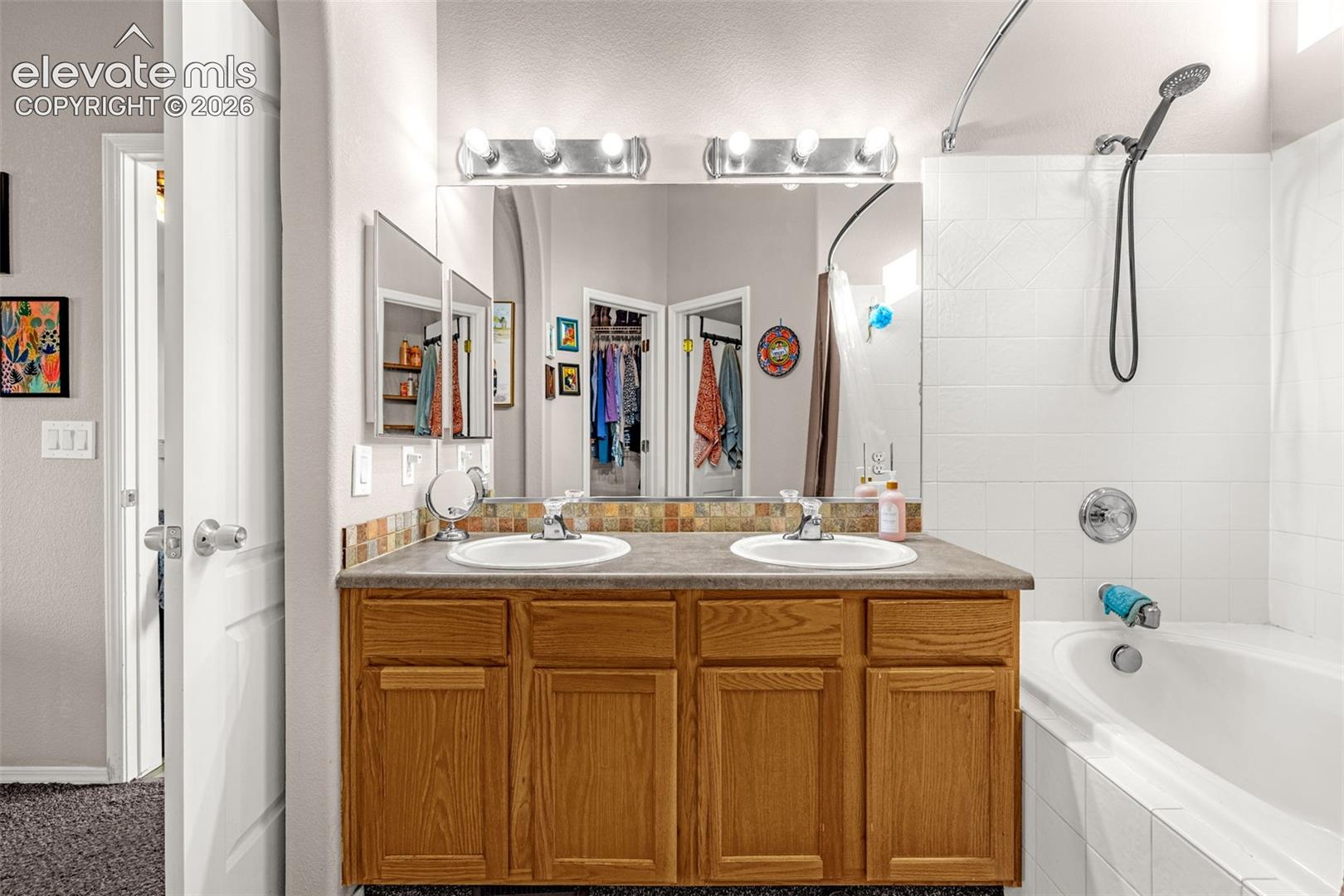 Bathroom featuring a dual-sink vanity with natural wood cabinetry, a decorative tiled backsplash, two integrated porcelain sinks, and a bathtub with a tiled surround