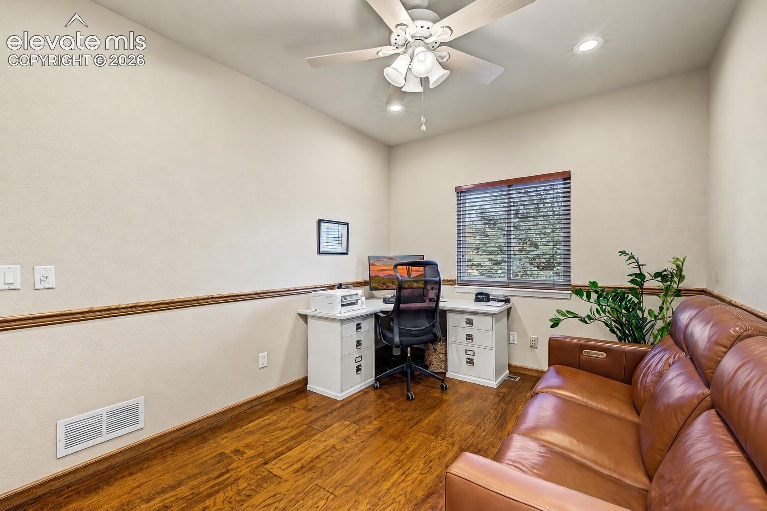 Additional view of the main-level bedroom currently used as a home office, featuring hardwood flooring, ceiling fan, recessed lighting, and a window overlooking mature trees.