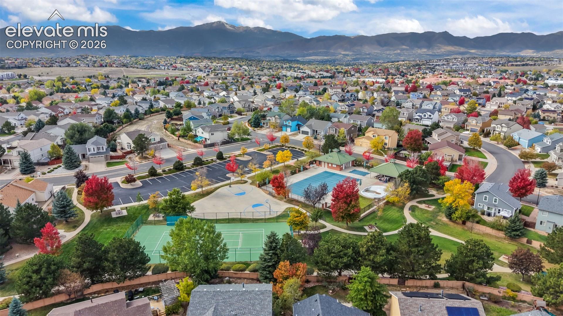 Aerial perspective of community amenities featuring a swimming pool, tennis court, dedicated parking area, and extensive green space set against a mountain backdrop