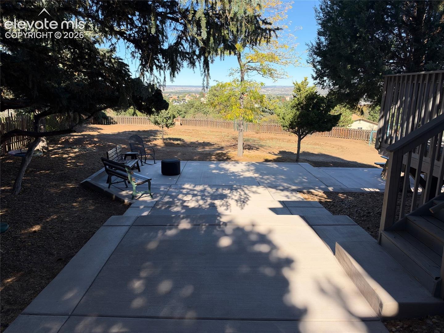 Multilevel concrete patio featuring a wooden staircase, perimeter wood-slat fencing, mature trees, and expansive terrain