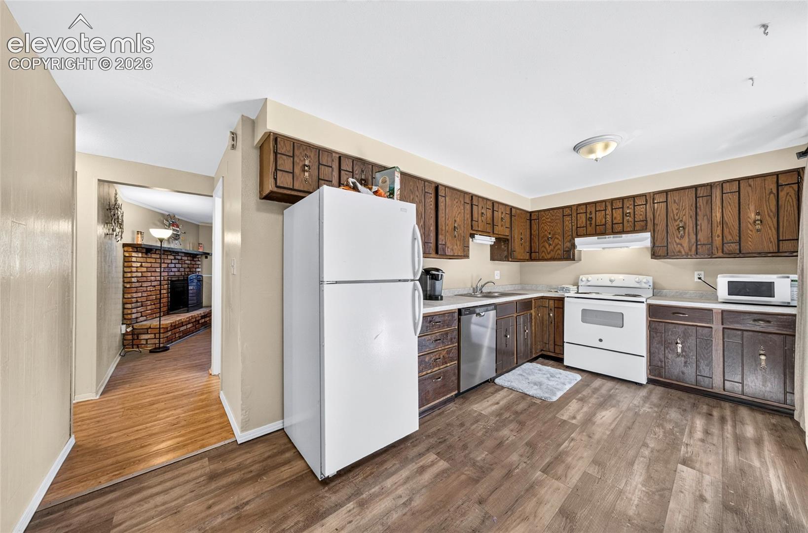 Kitchen featuring wood-finish cabinetry, stainless steel dishwasher, white range, wood-finish flooring, and a brick fireplace visible in the offset space