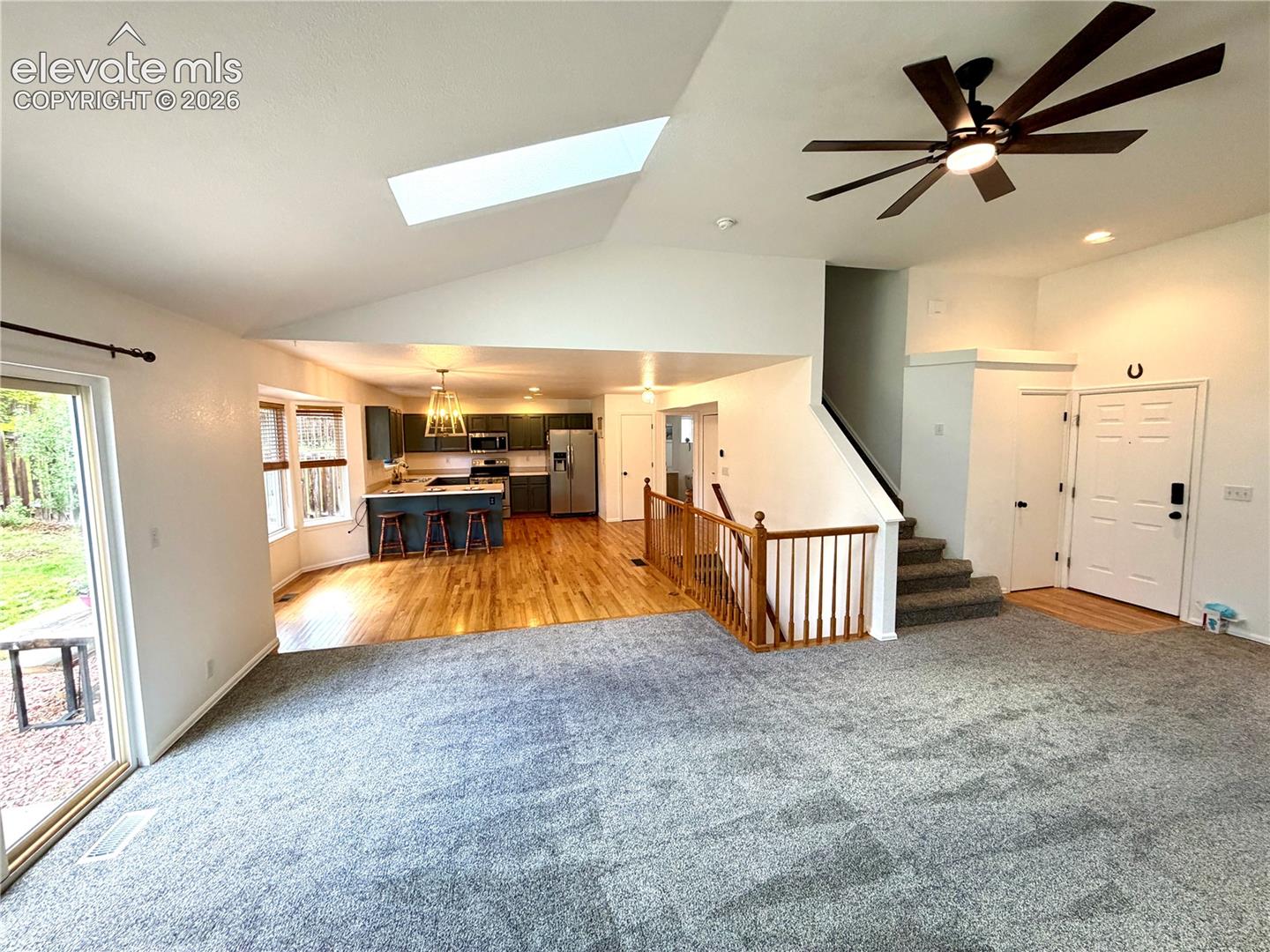 Open-concept living area featuring a vaulted ceiling with a skylight, a large ceiling fan with integrated lighting, and light gray carpeting