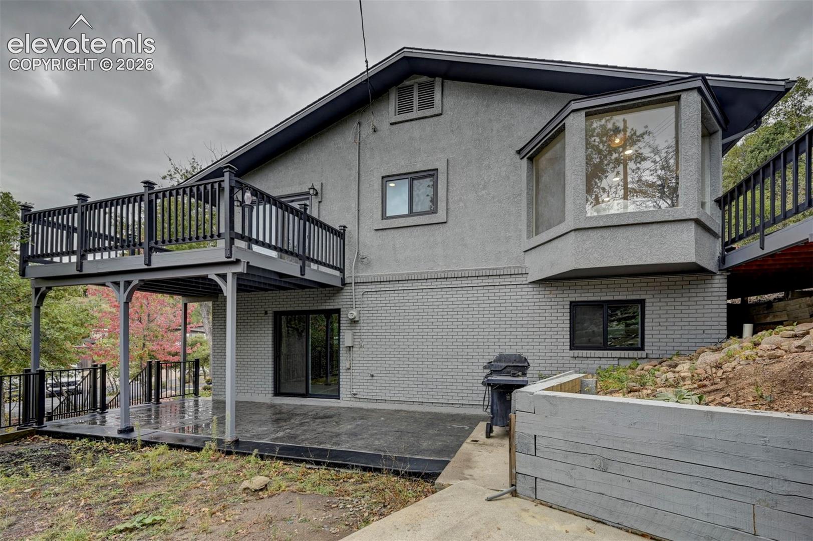 Stucco and painted brick exterior featuring an elevated deck with black railing, a paved ground-level patio, and a tiered timber retaining wall