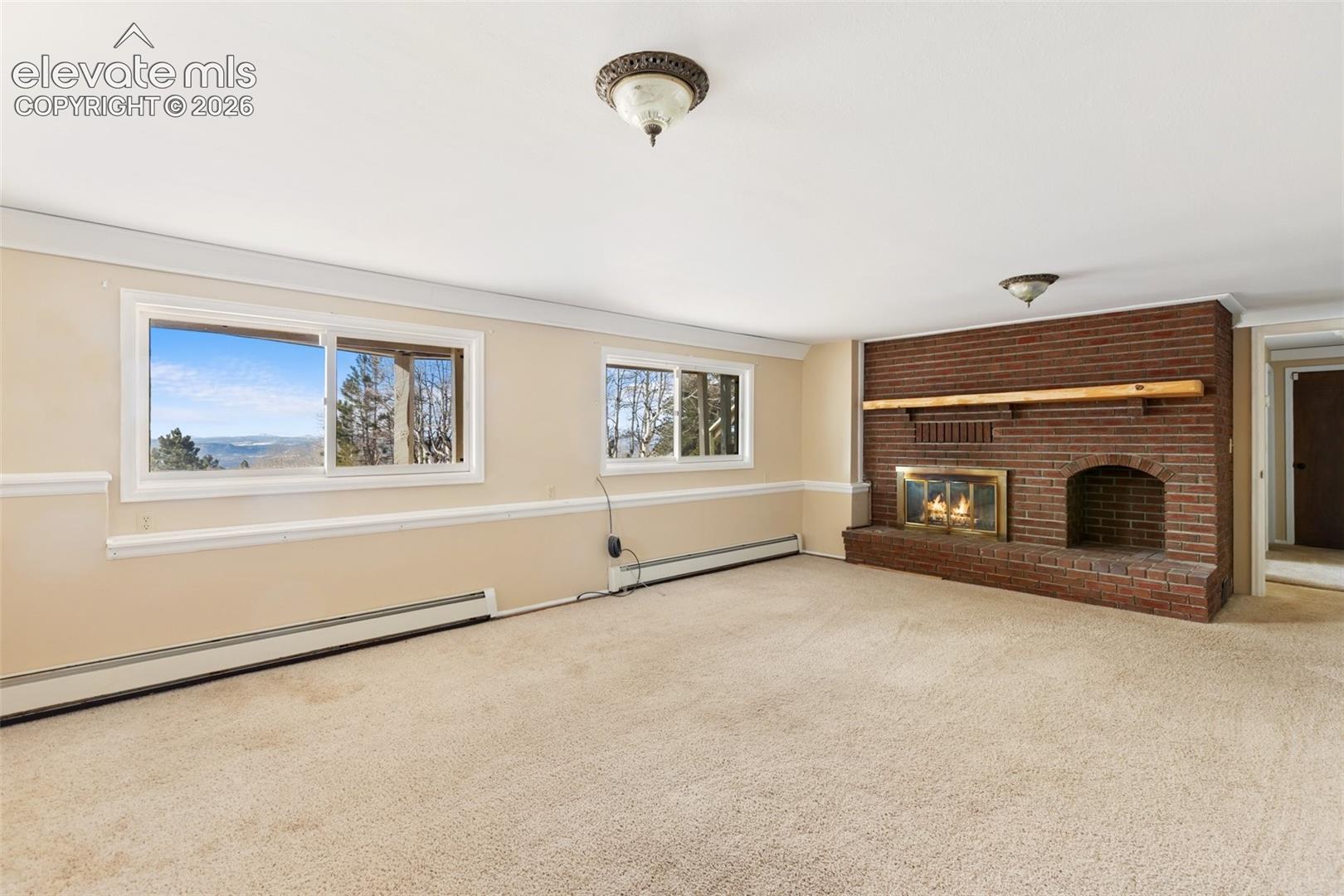 Expansive room featuring a red brick fireplace with a wood mantel, light-colored carpeting, two large windows, and a neutral wall color with chair rail molding