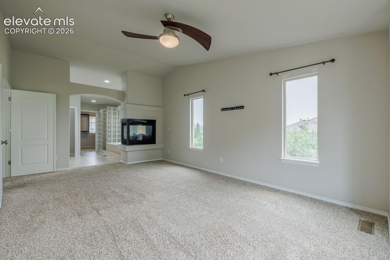 Carpeted main bedroom room featuring a ceiling fan with light fixture, two tall windows, and a built-in fireplace with a stone-finish hearth