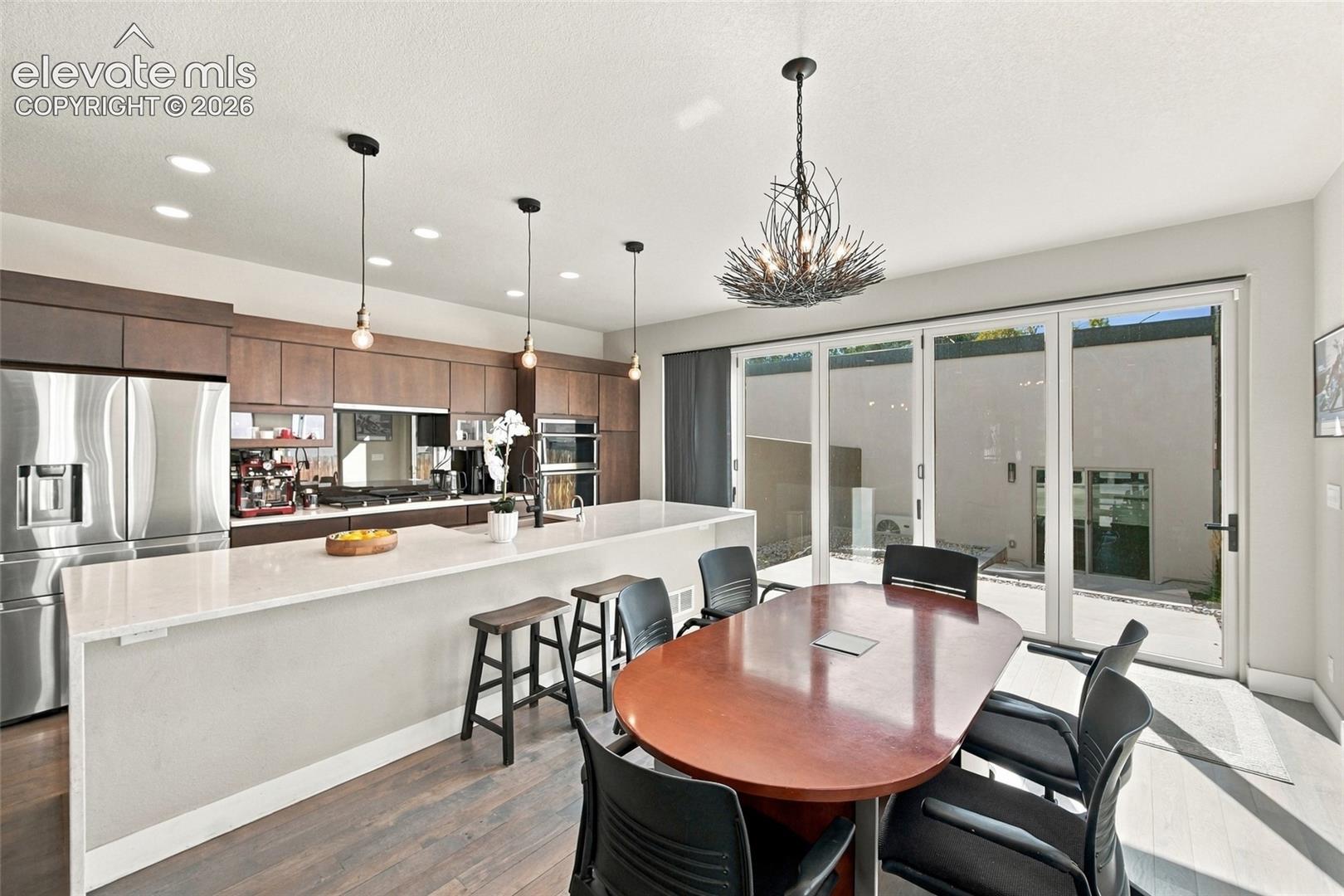 Modern kitchen featuring flat-panel cabinetry, a spacious island with a light-toned countertop, integrated wall ovens, stainless steel appliances, and oversized glass sliding accordion doors that open up for indoor outdoor living